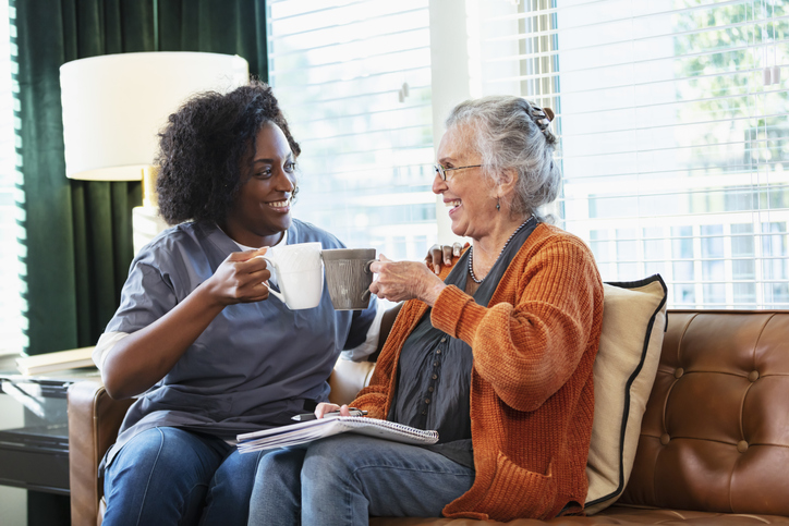 A senior woman in her 80s sitting on the living room couch, having coffee and conversing with a home healthcare aide, an African-American woman in her 30s