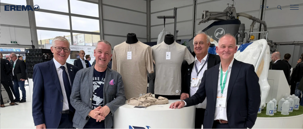 Manfred Hackl (EREMA Group), Graham Ross (BlockTexx®), Wolfgang Hermann (EREMA Group) und Adrian Jones (BlockTexx®) neben T-Shirts aus 100% recyceltem Polyester im Edvanced Recycling Center, wo EREMA während der K-Messe Live-Recycling und Produkte aus Rezyklaten vorstellt. 