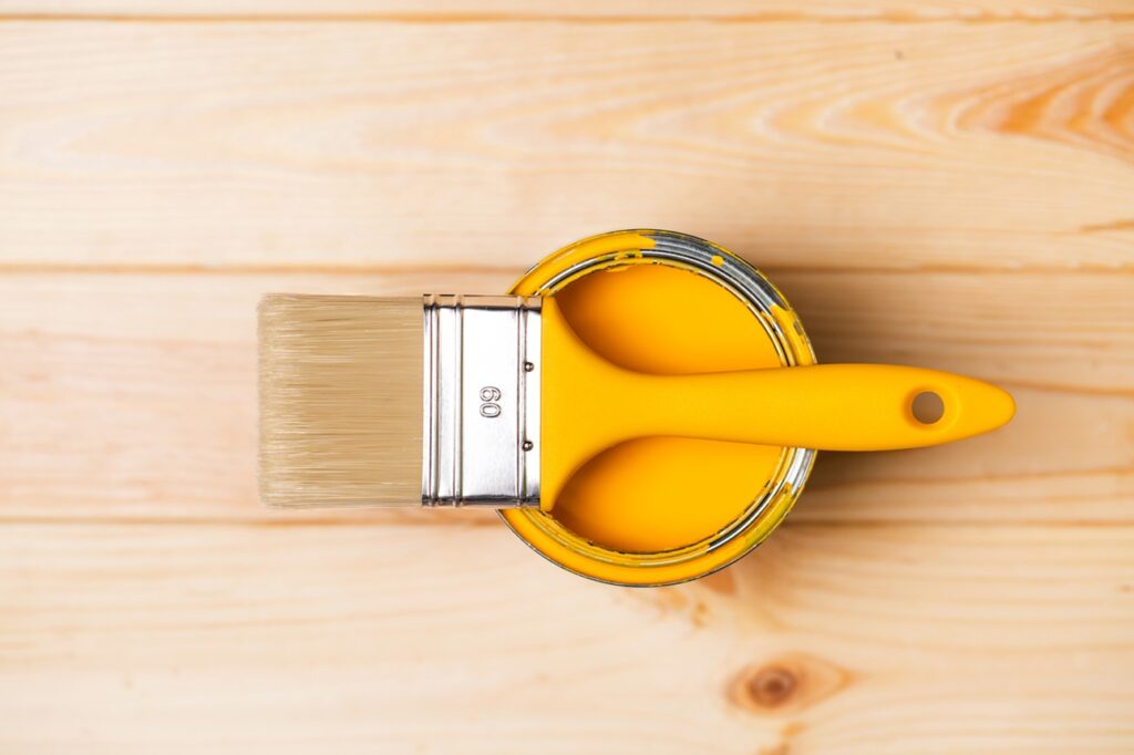 Top view of clean paintbrush placed on top of open metal bucket with liquid pigment on wooden background